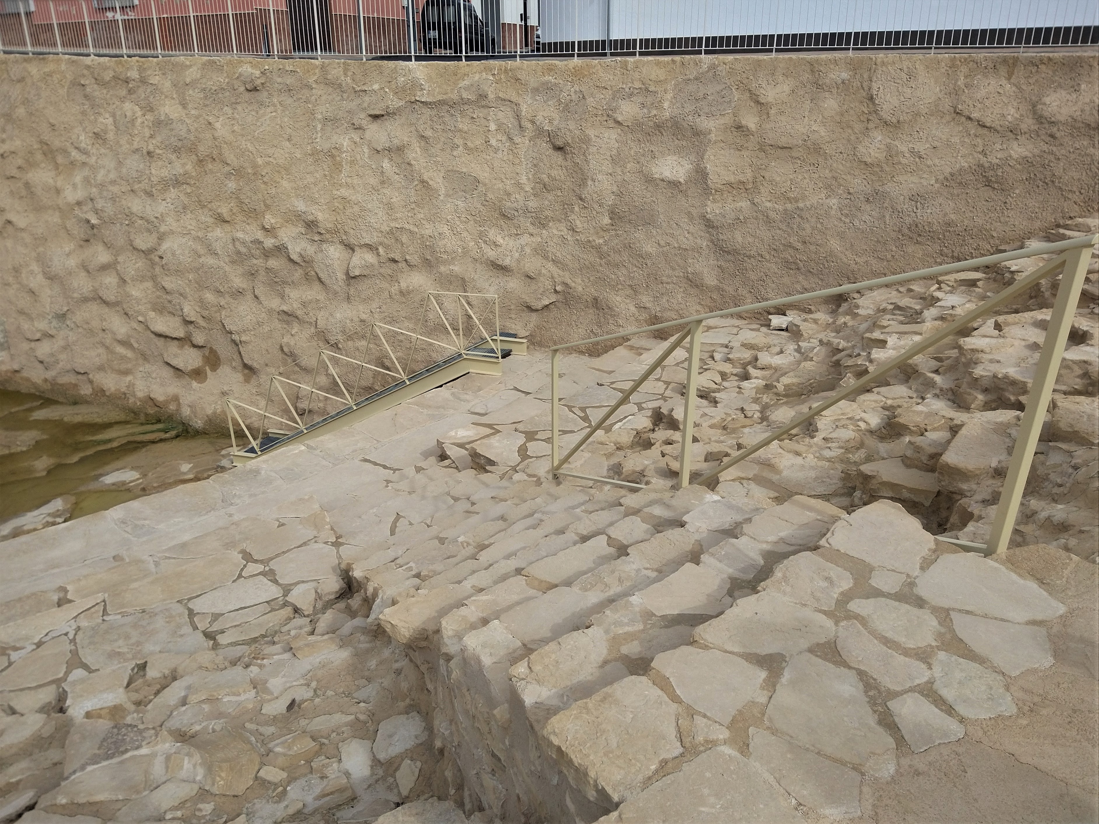Vista parcial de la cavea. Abajo la arena parcialmente inundada debido a las lluvias. Al fondo la pared de tierra que impide la continuación de los trabajos de excavación, ya que sobre la gran acumulación de sedimentos se pueden apreciar las viviendas unifamiliares existentes.